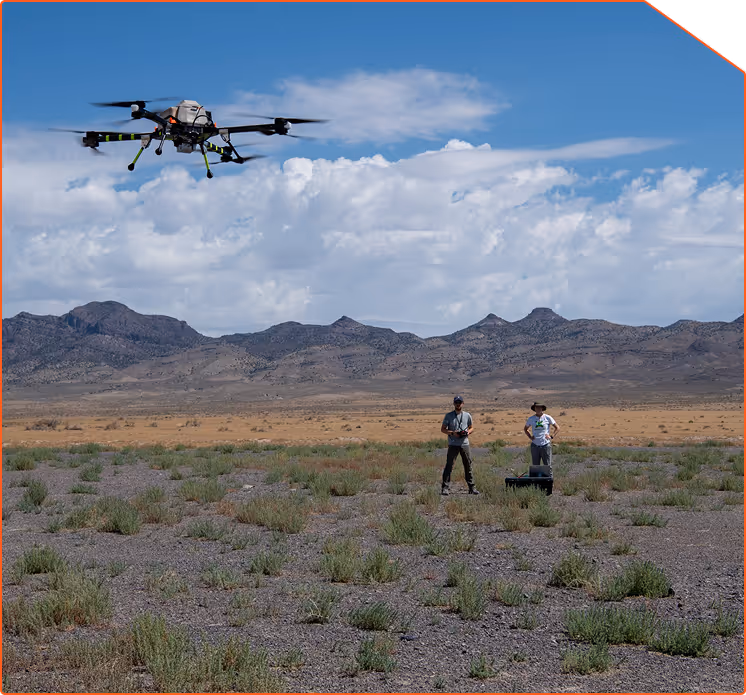 Two people operating a large drone flying over a rocky desert landscape with mountains and a cloudy sky in the background.