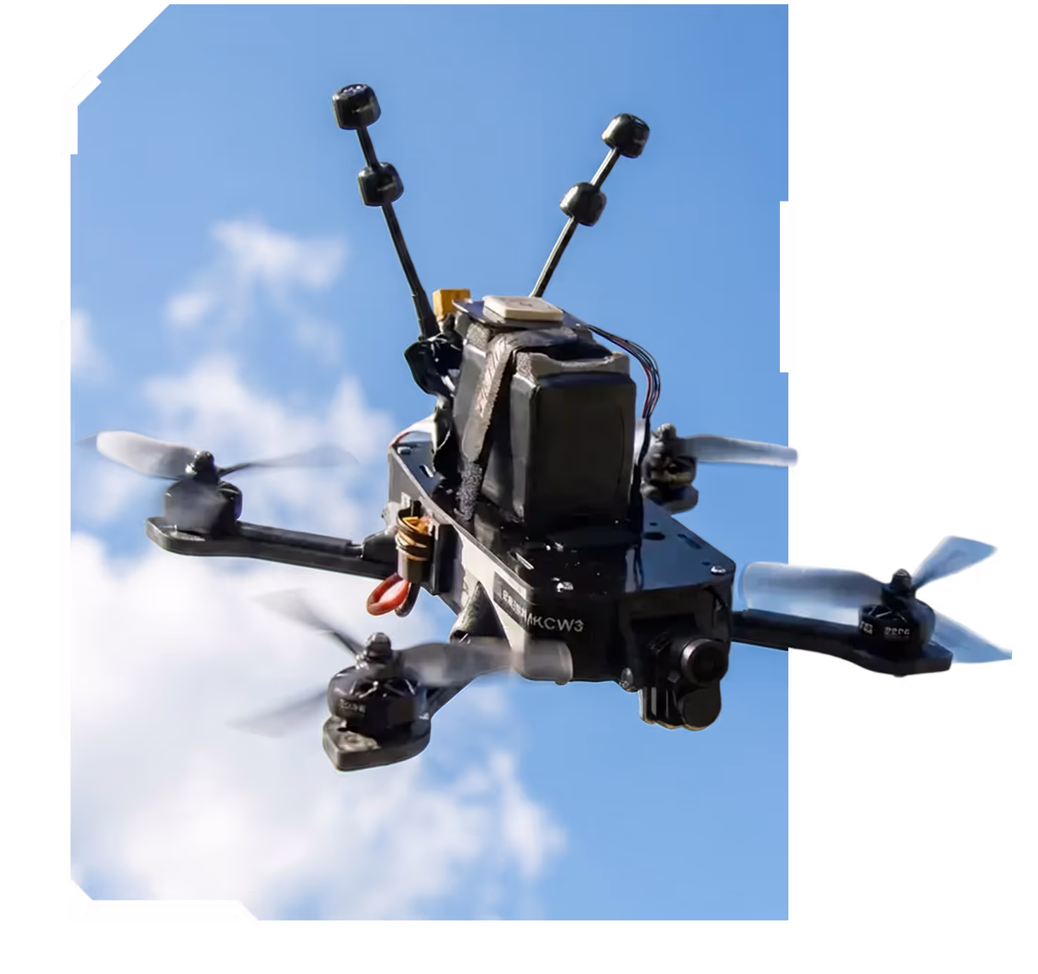 Black quadcopter drone flying against a blue sky with clouds, featuring four spinning propellers and dual antennas.