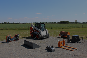 Compact skid-steer loader surrounded by various interchangeable attachments on gravel with a field and blue sky in the background.