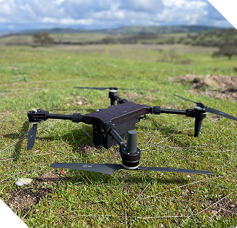 Black quadcopter drone resting on green grass with a cloudy sky and distant hills in the background.
