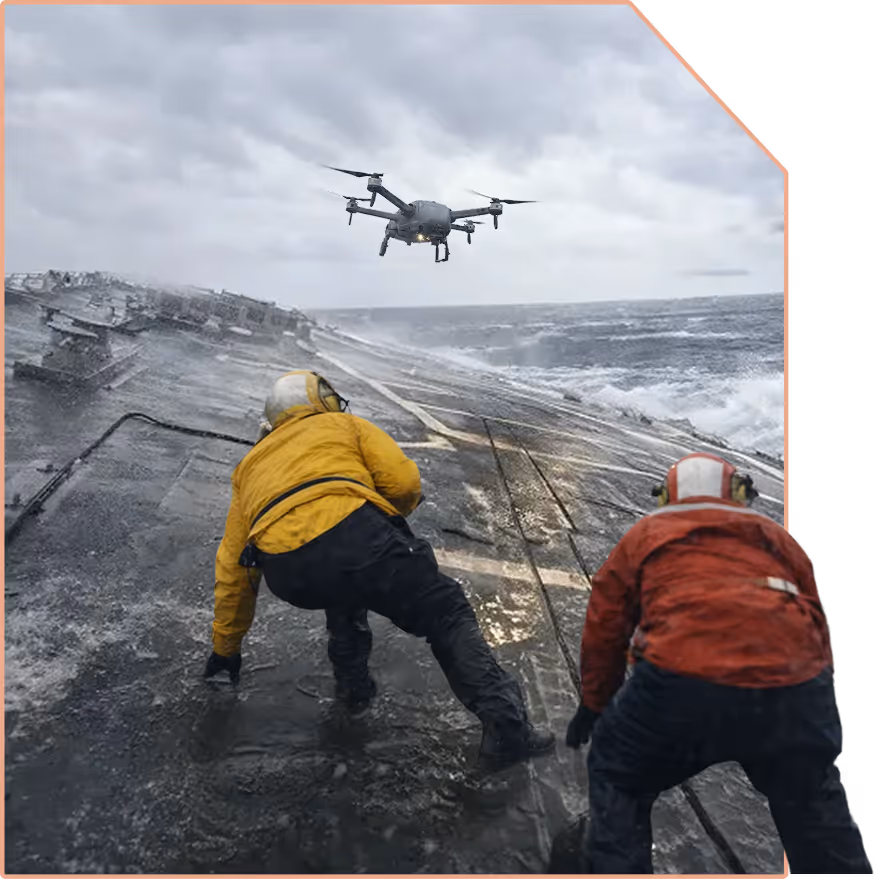 Two crew members in yellow and red jackets crouch on a wet aircraft carrier deck during rough seas as a drone hovers nearby.