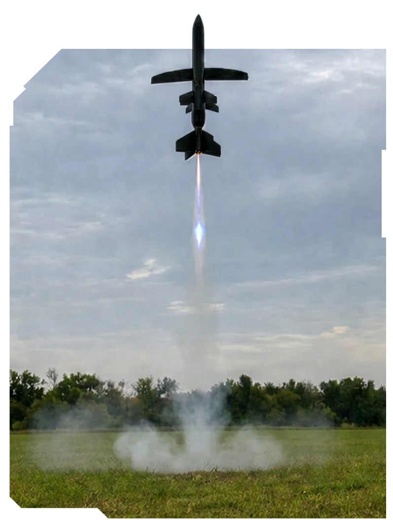 Rocket launching vertically from a grassy field with trees and a cloudy sky in the background.