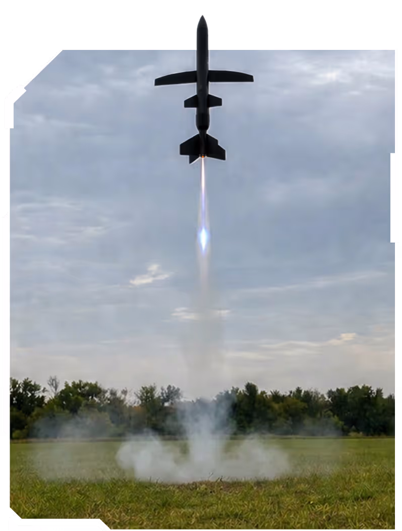 Rocket launching vertically from a grassy field with trees and a cloudy sky in the background.