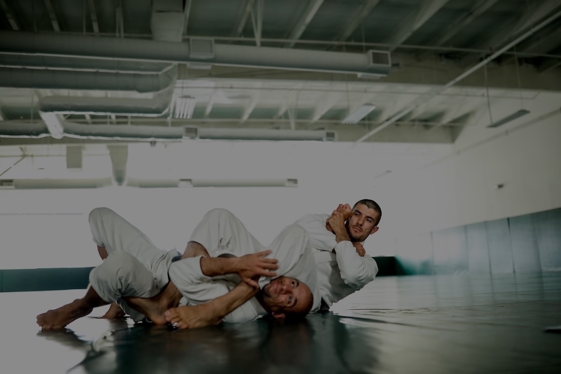 Two men in white gis practicing Brazilian Jiu-Jitsu on a black mat, one applying a straight armlock from mount position.