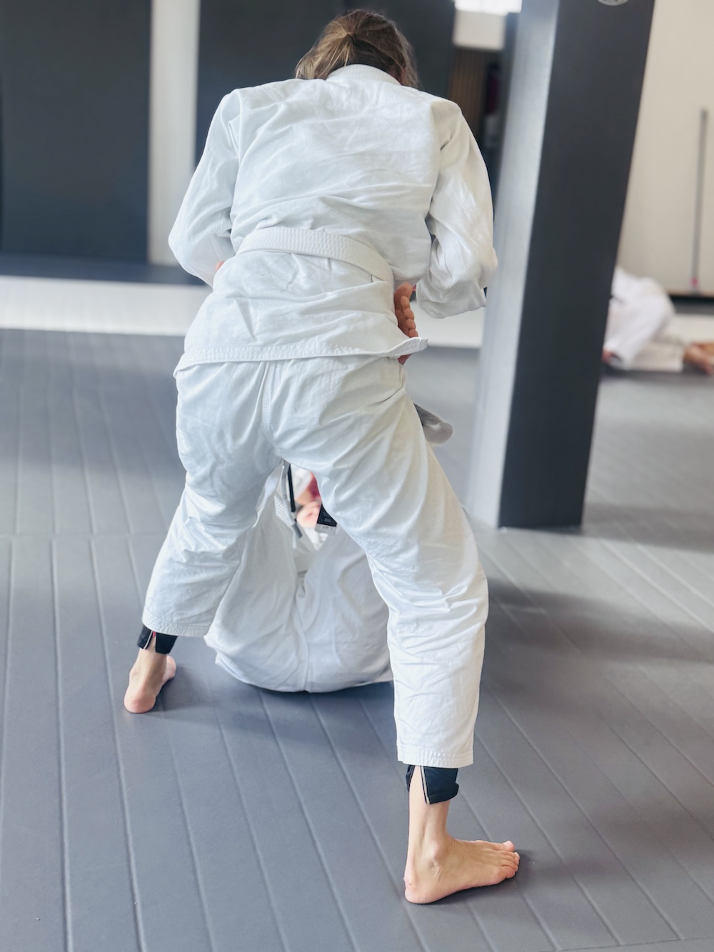 Bright martial arts gym with black and white padded flooring, gymnastic rings hanging from the ceiling, and windows letting in natural light.