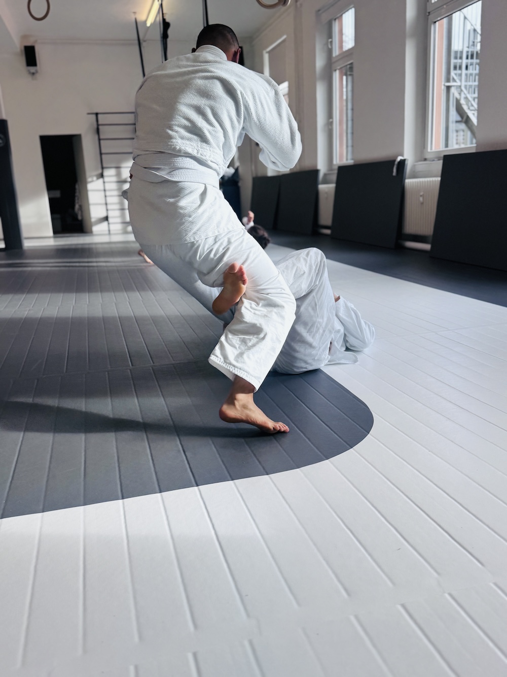 Two pairs of people practicing Brazilian Jiu-Jitsu on mats in a gym, wearing white gi uniforms.