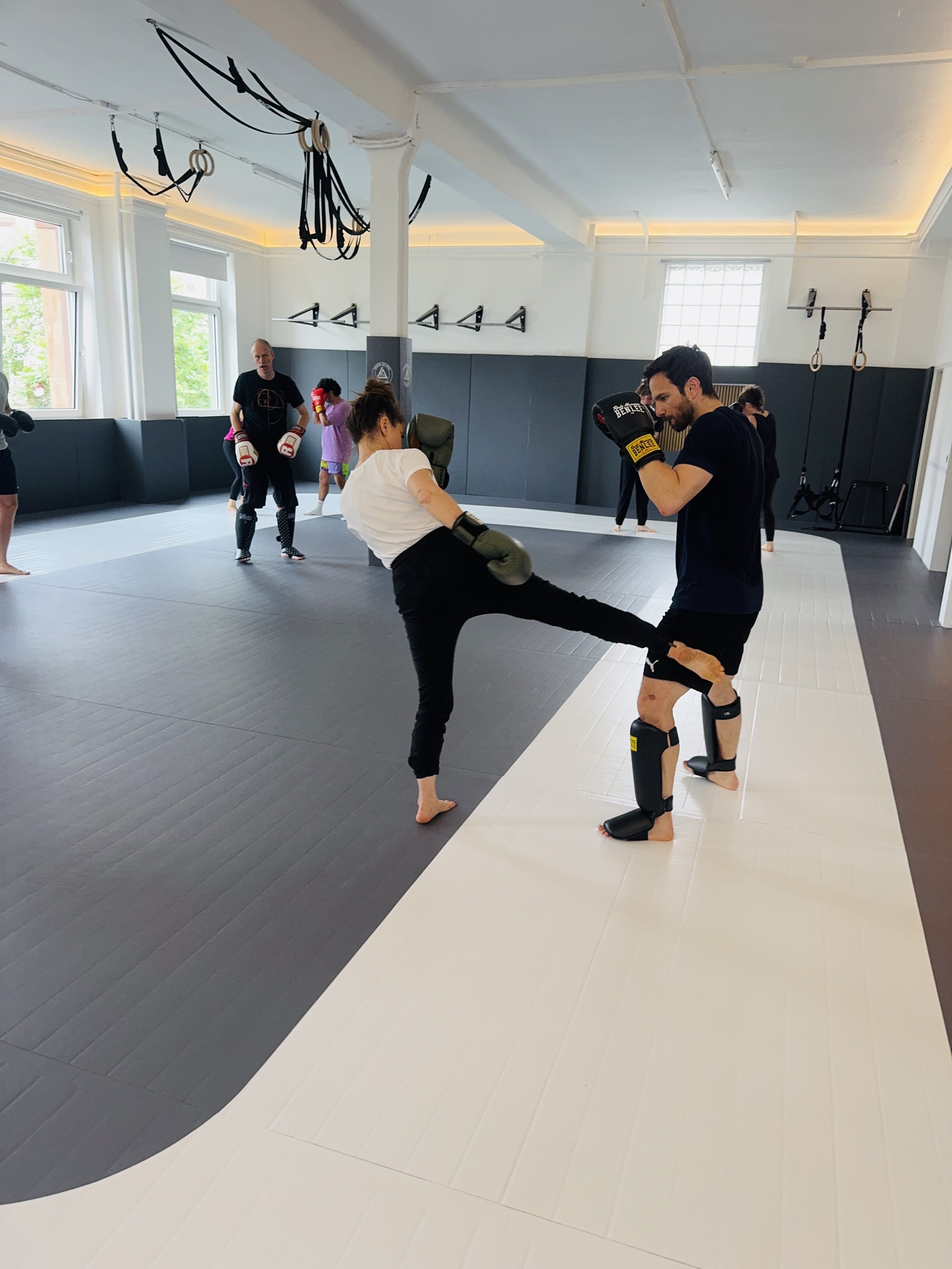 Group of six people standing in a line on martial arts mats wearing white Brazilian Jiu-Jitsu gis with arms crossed.