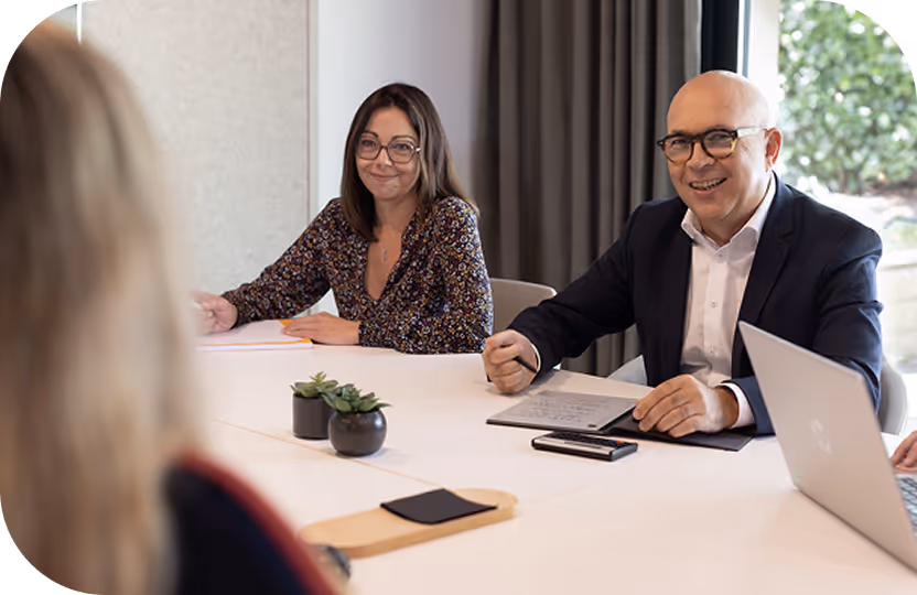 Deux professionnels en réunion, une femme souriante et un homme portant des lunettes, assis à une table blanche avec un ordinateur portable et des documents.