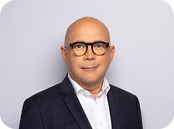 Portrait d'un homme chauve portant des lunettes et un costume noir avec une chemise blanche, devant un fond gris clair.