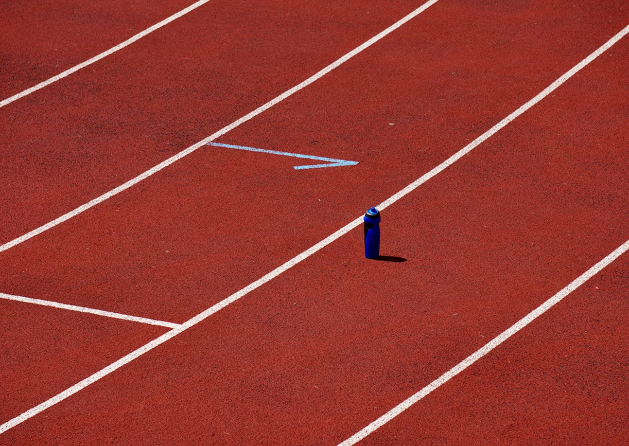 Blue water bottle standing on an empty red running track with white lane markings and a blue arrow pointing forward.