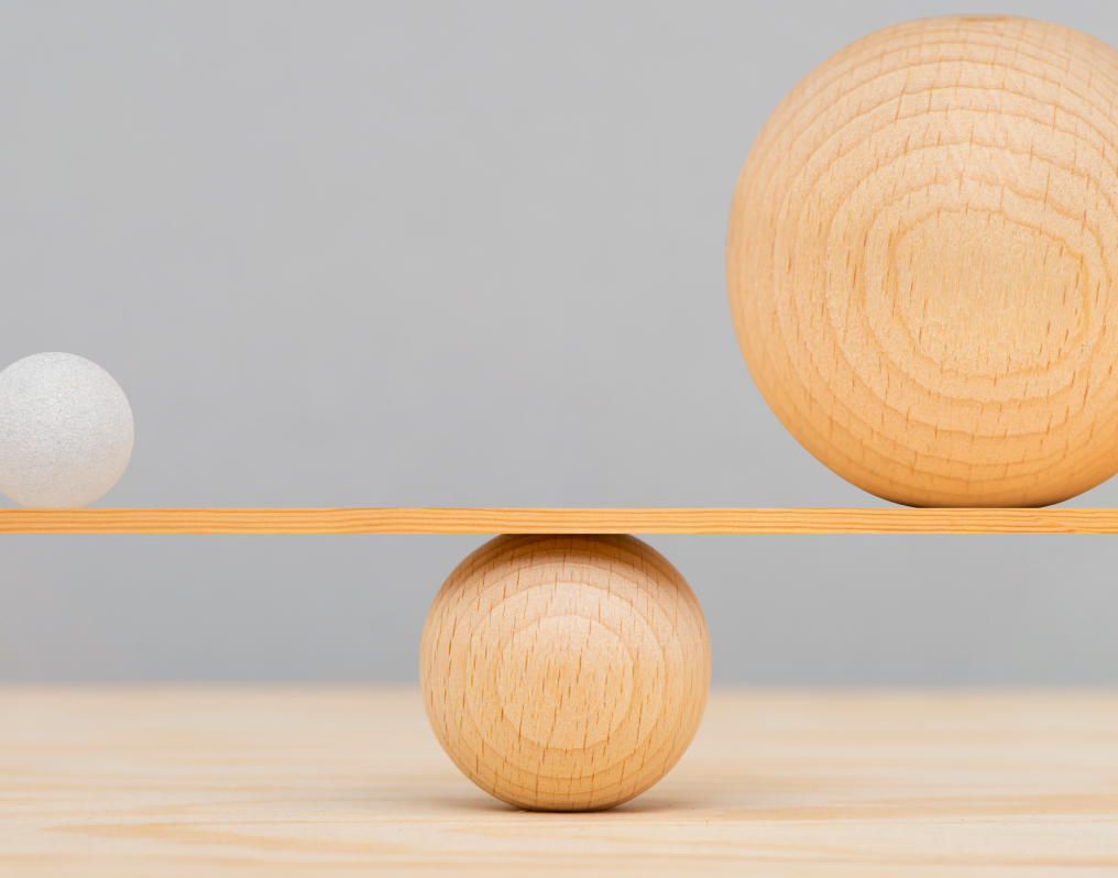 A large wooden sphere and a small white sphere balanced on opposite ends of a wooden plank resting on a medium-sized wooden sphere.