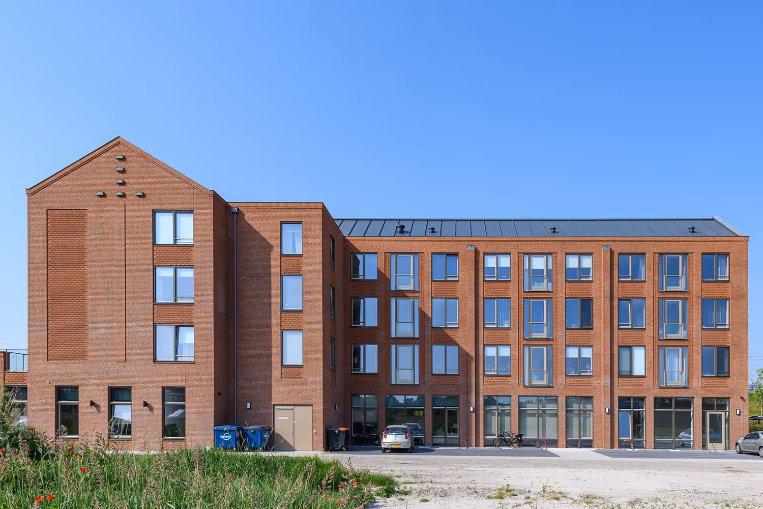 Modern appartementsgebouw met rode bakstenen, grote ramen en een metalen dak onder een heldere blauwe lucht.