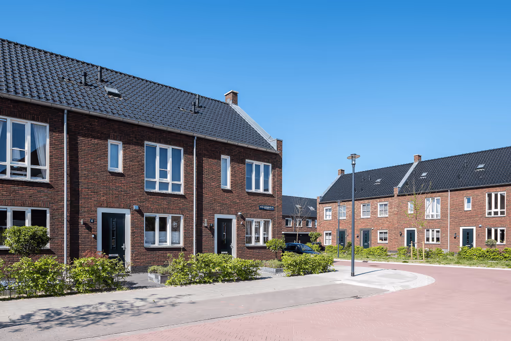 Moderne rode bakstenen rijtjeshuizen met puinversterkte zwarte daken en groenblijvende struiken bij een rustige straat onder een heldere blauwe hemel.