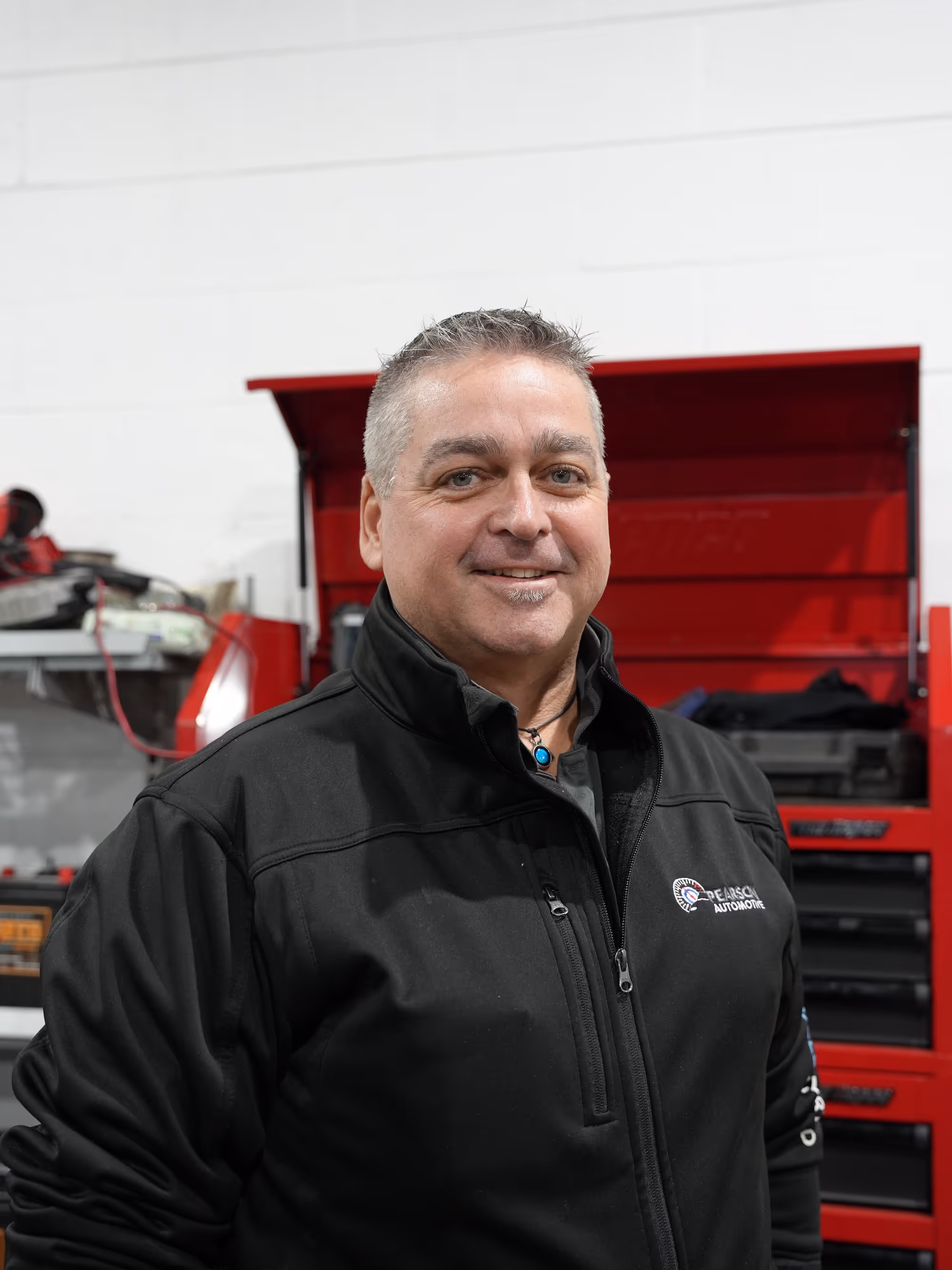 A friendly mechanic from the Pearson Automotive team in Milton, wearing a branded jacket and standing in front of a red tool chest in the workshop.