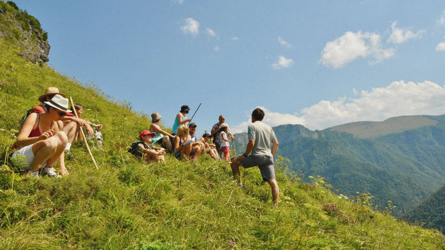 Groupe de randonneurs assis sur une pente herbeuse en montagne, avec un guide debout expliquant quelque chose.