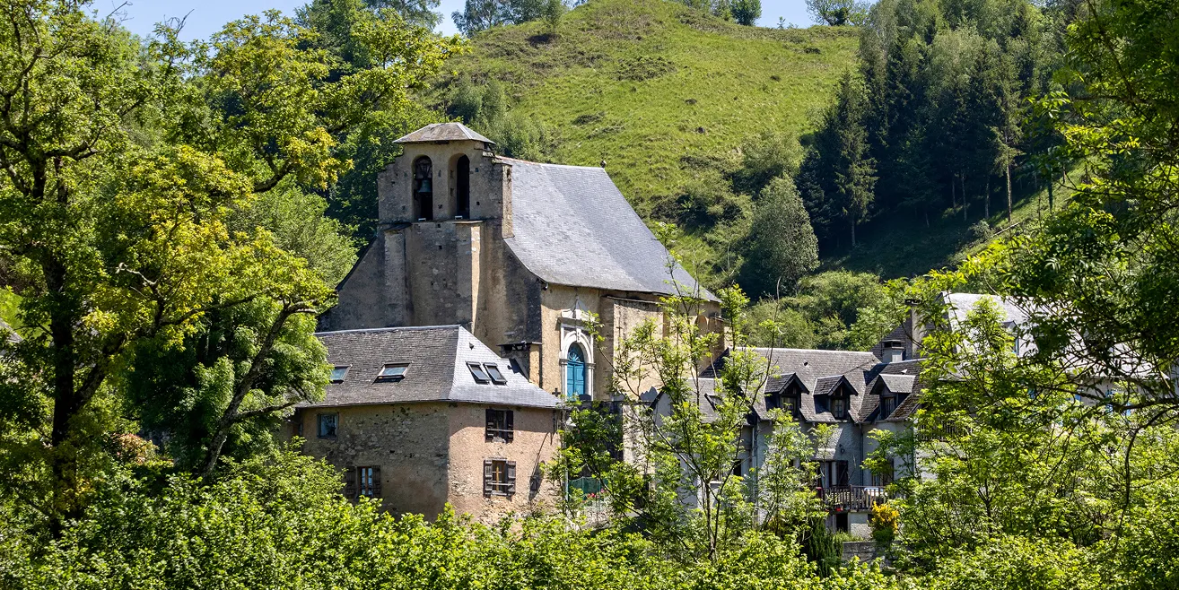 Vieilles maisons en pierre avec un clocher sous un ciel bleu, entourées d'arbres verts et d'une colline herbeuse.