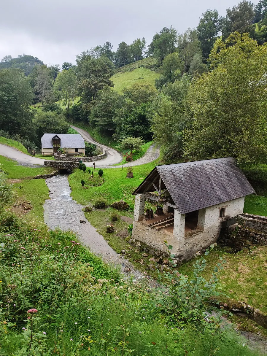 Petits bâtiments en pierre près d'un ruisseau dans une vallée verte et boisée.