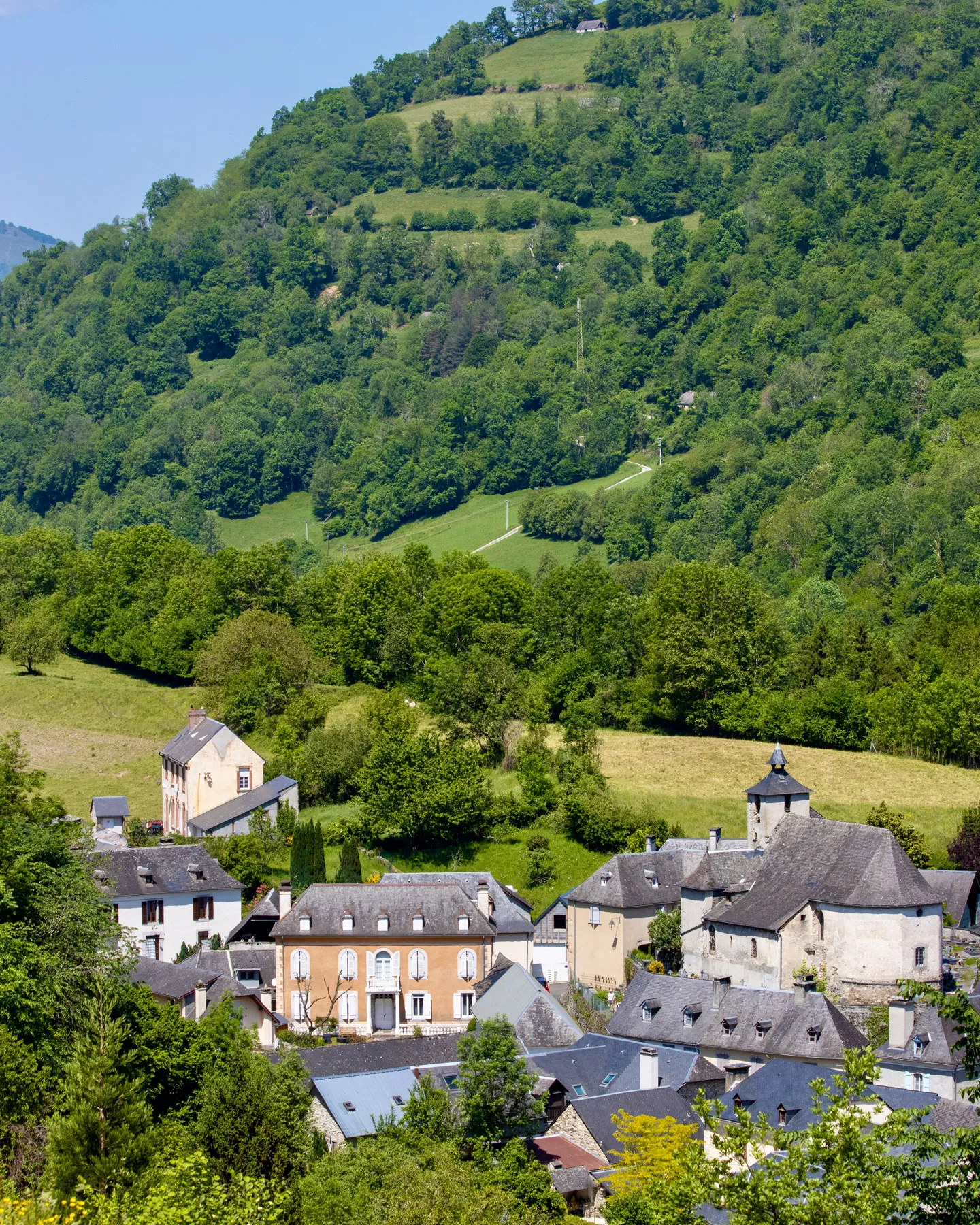Village nestled at the base of lush green hills with a church and traditional houses under clear blue sky.