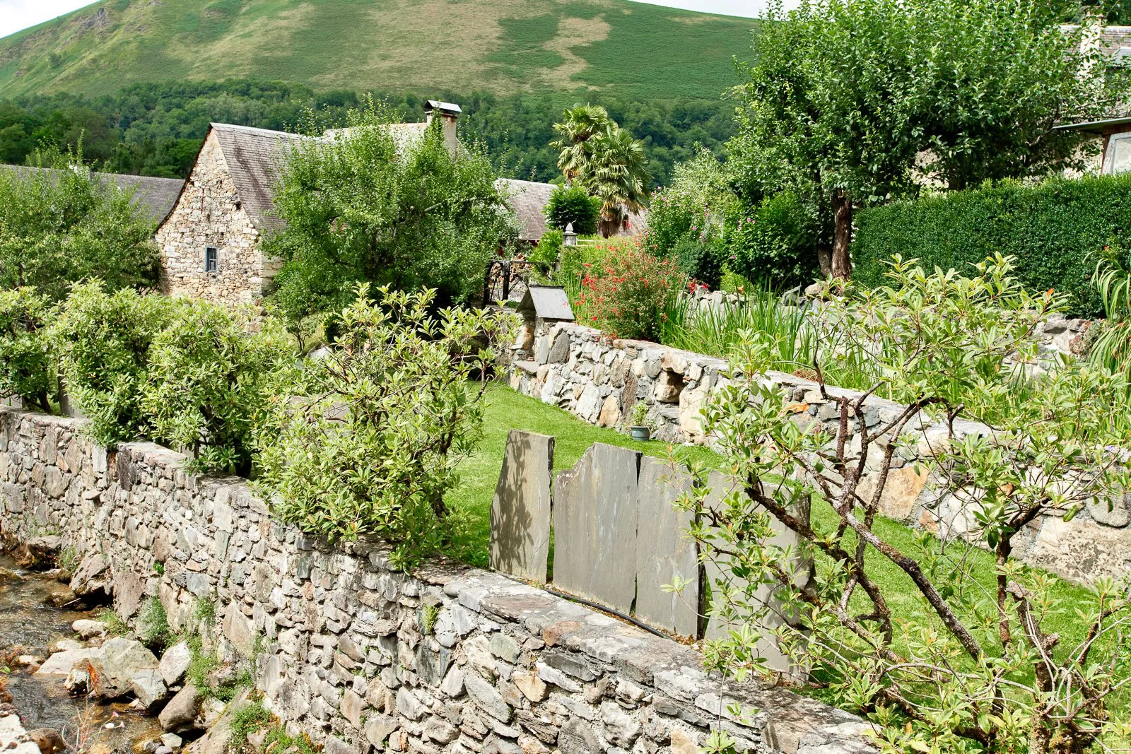Jardin verdoyant avec murs en pierre, arbres, maison en pierre et colline herbeuse en arrière-plan.