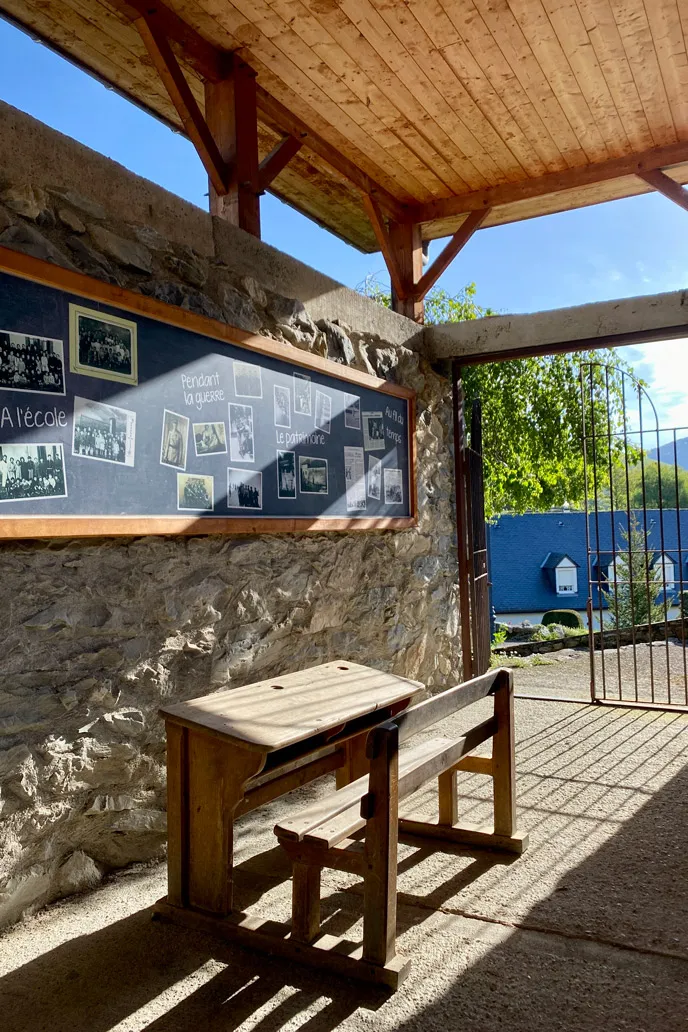 Ancien bureau d'école en bois sous un toit en bois, près d'un panneau mural avec photos historiques.