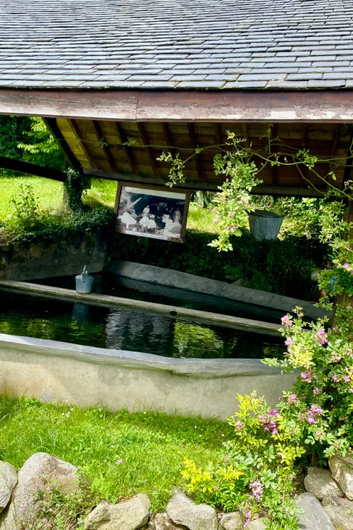 Lavoir en pierre avec eau, abrité par un toit en tuiles, entouré de verdure et fleurs.