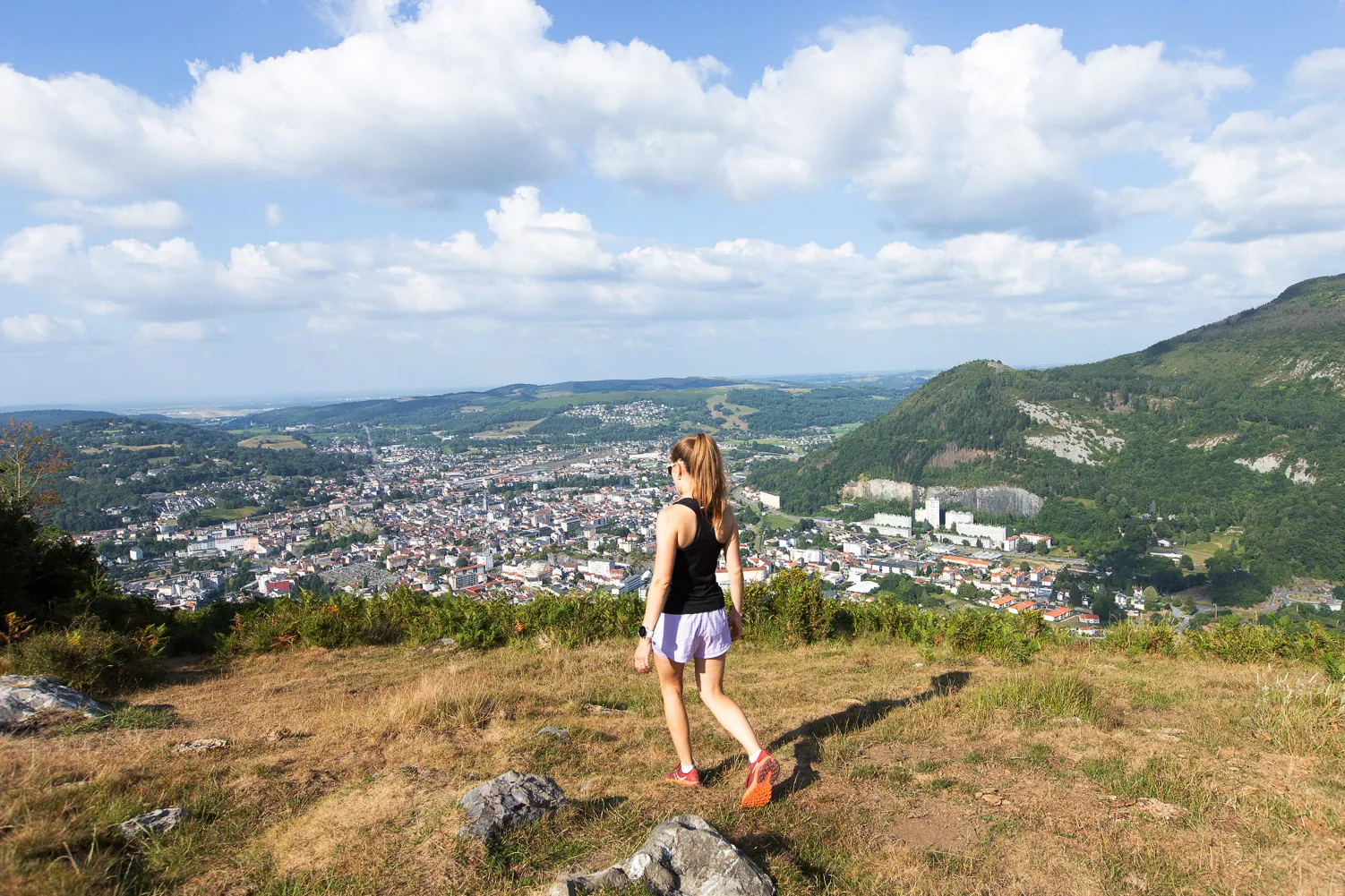 Une femme marche dans une clairière avec vue panoramique sur une ville enclavée entre collines verdoyantes.
