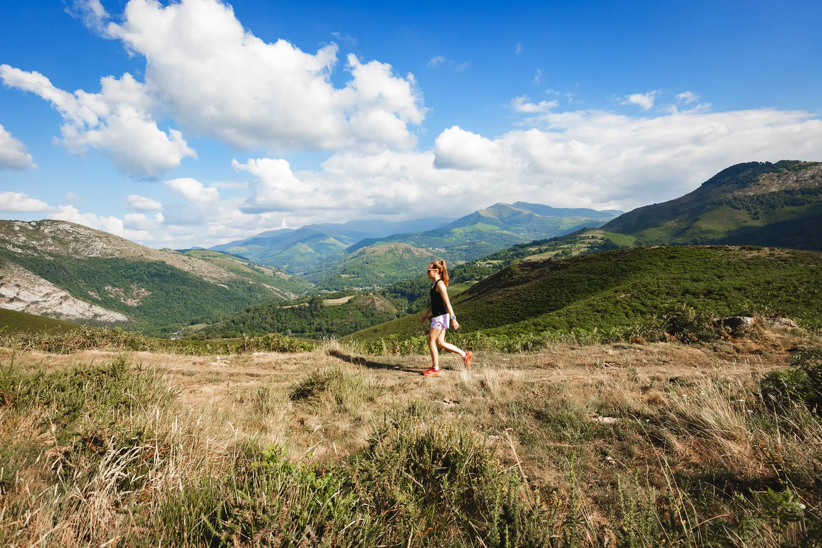 Jeune femme marchant sur un sentier en montagne sous un ciel bleu avec des nuages blancs.