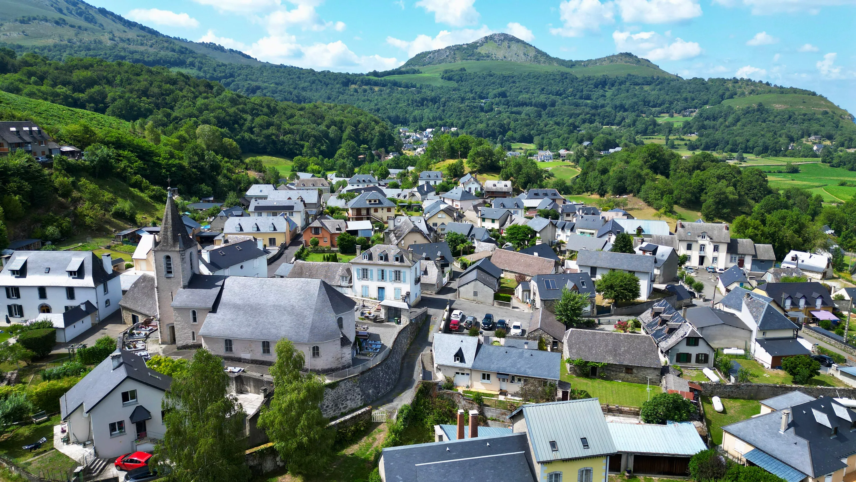 Village de montagne avec maisons grises, une église au toit pointu, entouré de collines verdoyantes.
