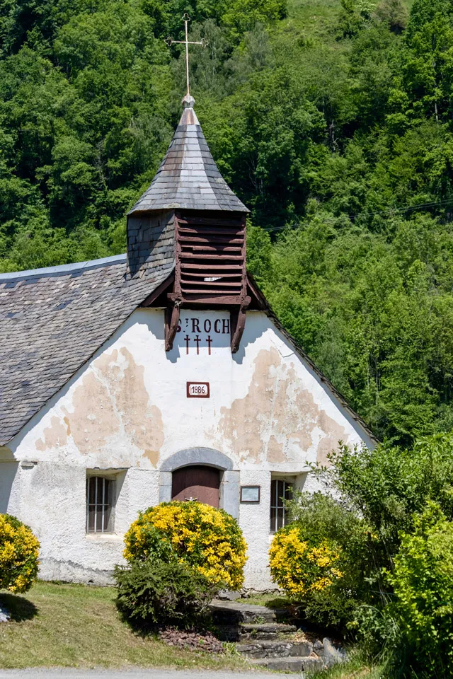 Chapelle Saint-Roch blanche avec toit en ardoise, entourée d'arbustes jaunes et de forêt verte.