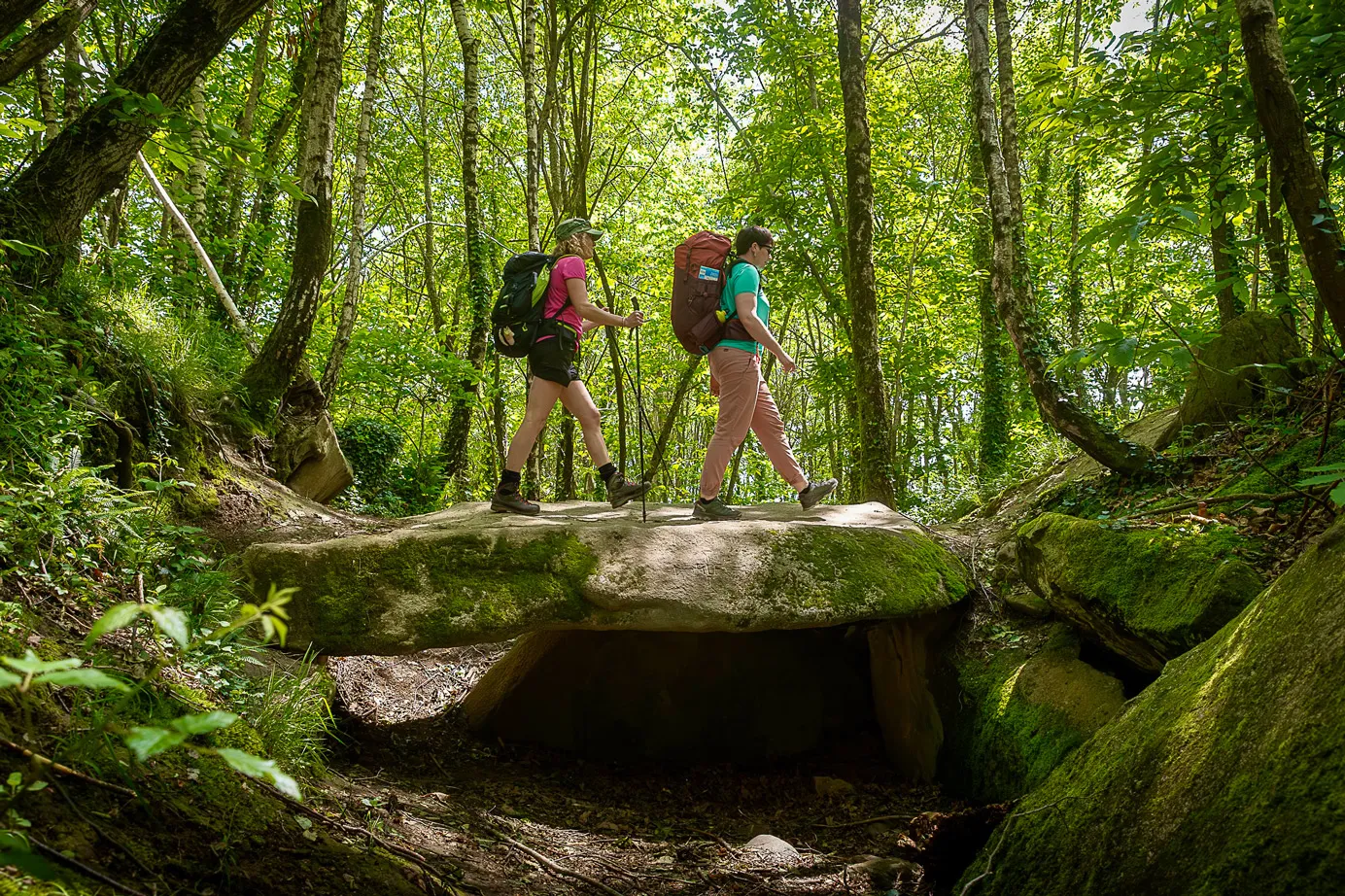 Deux randonneurs traversant un dolmen recouvert de mousse en forêt lumineuse.