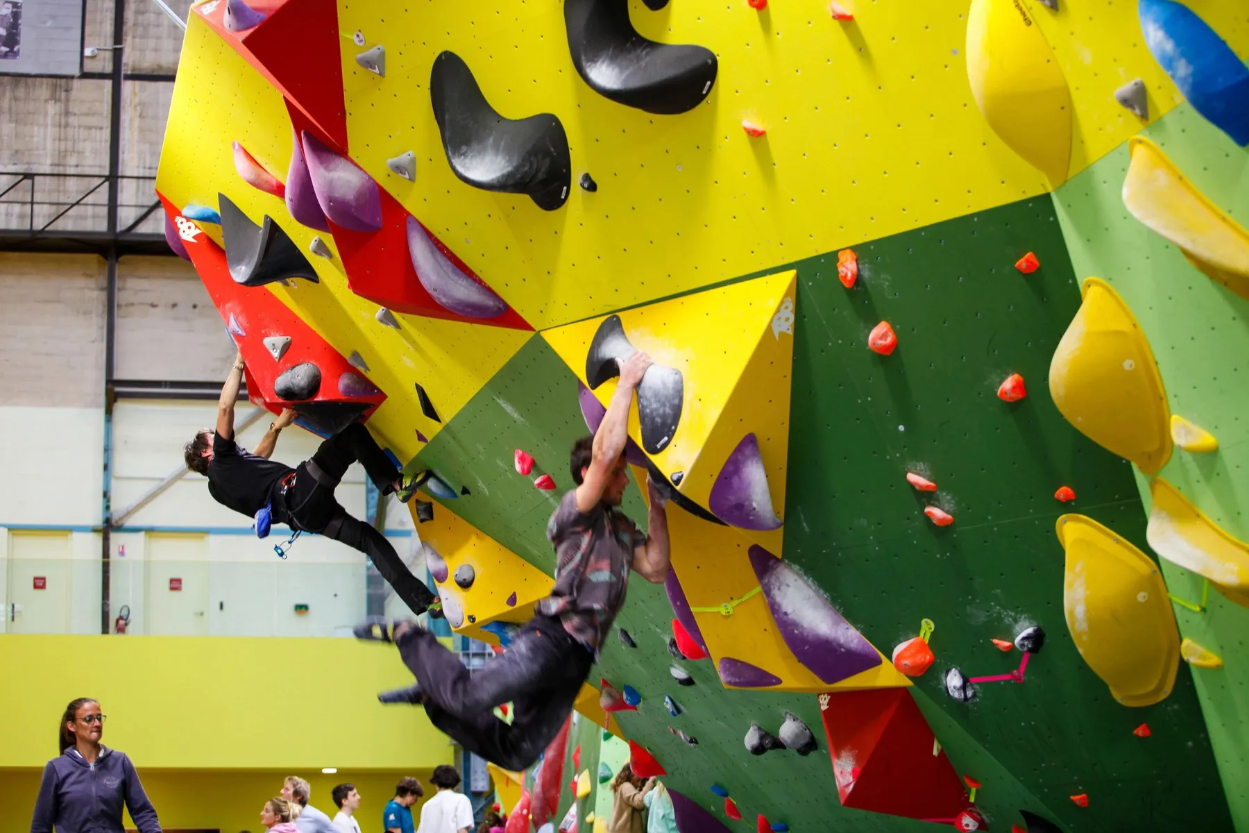 Deux grimpeurs escaladent un mur d'escalade coloré avec des prises variées en salle.