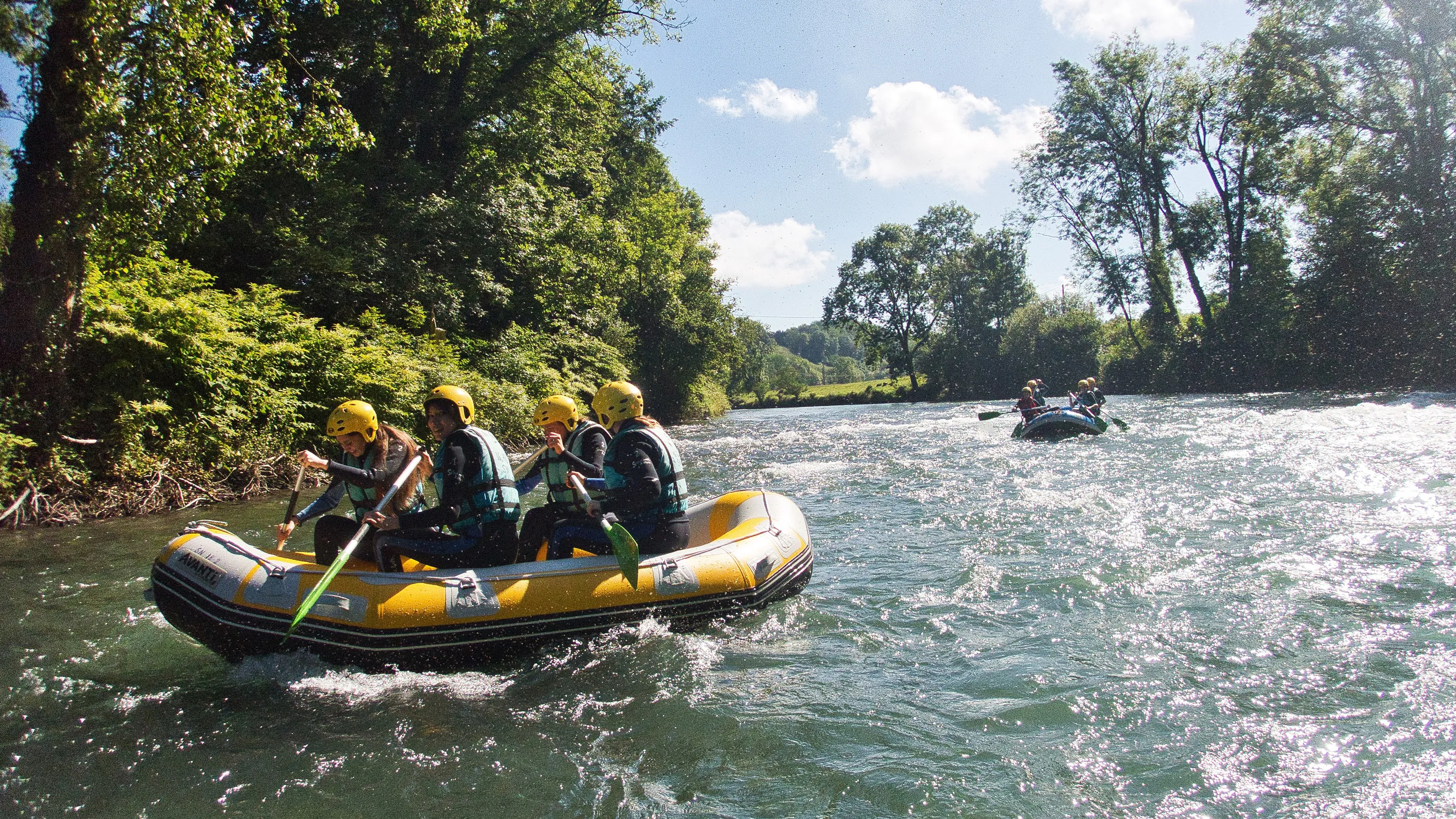 Deux équipes en raft jaune et bleu pagayant sur une rivière agitée bordée d'arbres verts.