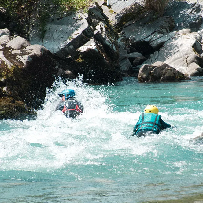 Deux personnes avec casques s'engagent dans une eau vive turquoise entre des rochers.