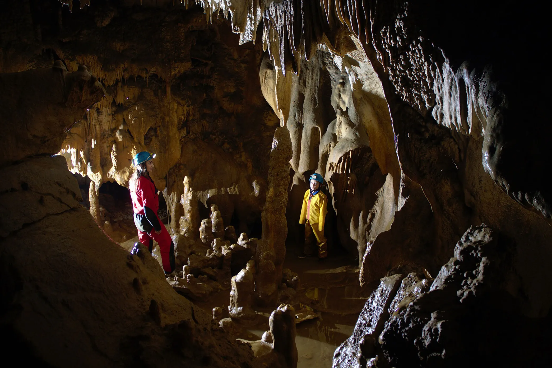 Deux spéléologues en combinaison avec casque et lampe explorent une grotte avec stalactites et stalagmites.