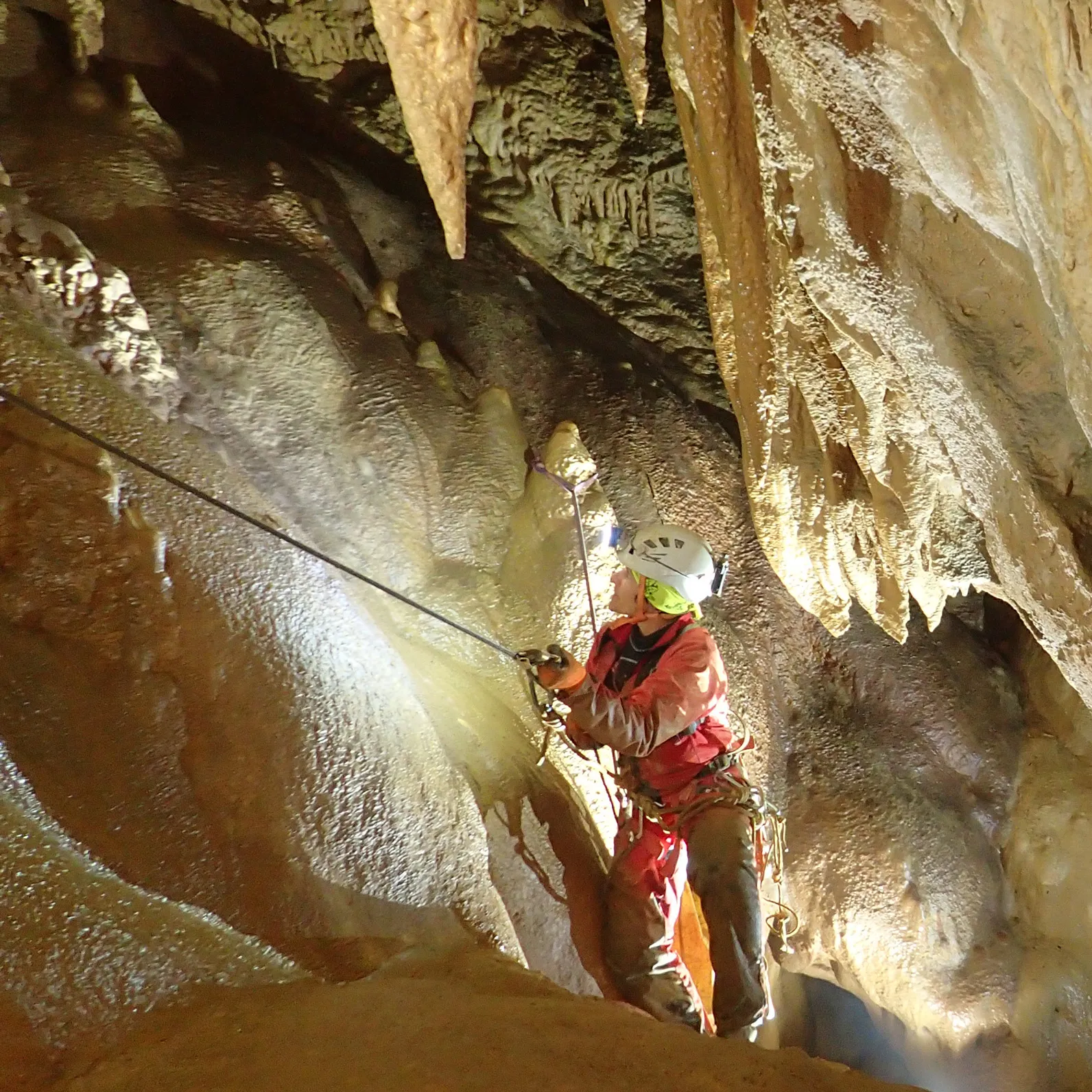 Spéléologue équipé descend dans une grotte éclairée avec stalactites et parois rocheuses humides.