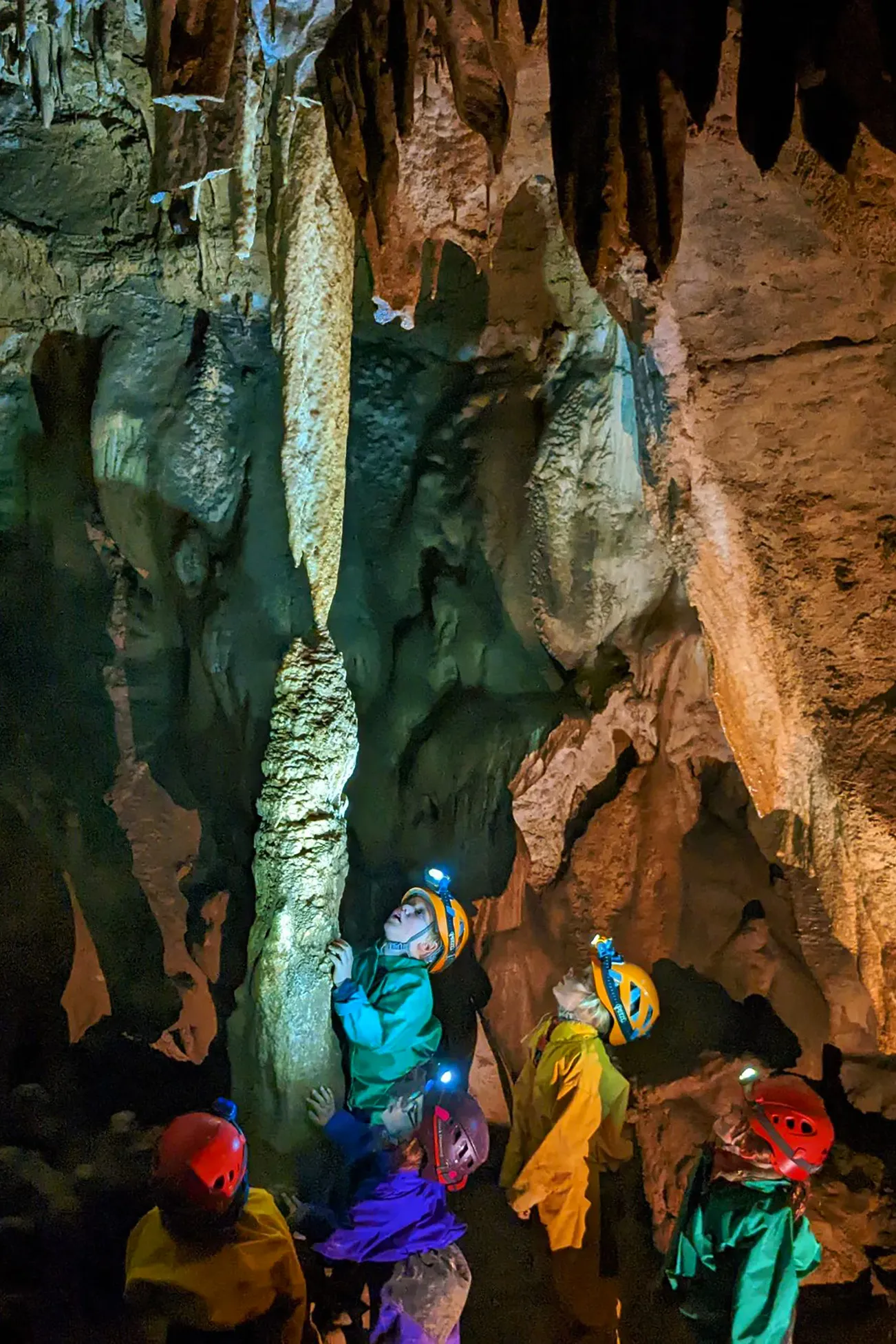 Enfants en combinaison et casque avec lampe explorant une grotte avec stalactites et stalagmites.