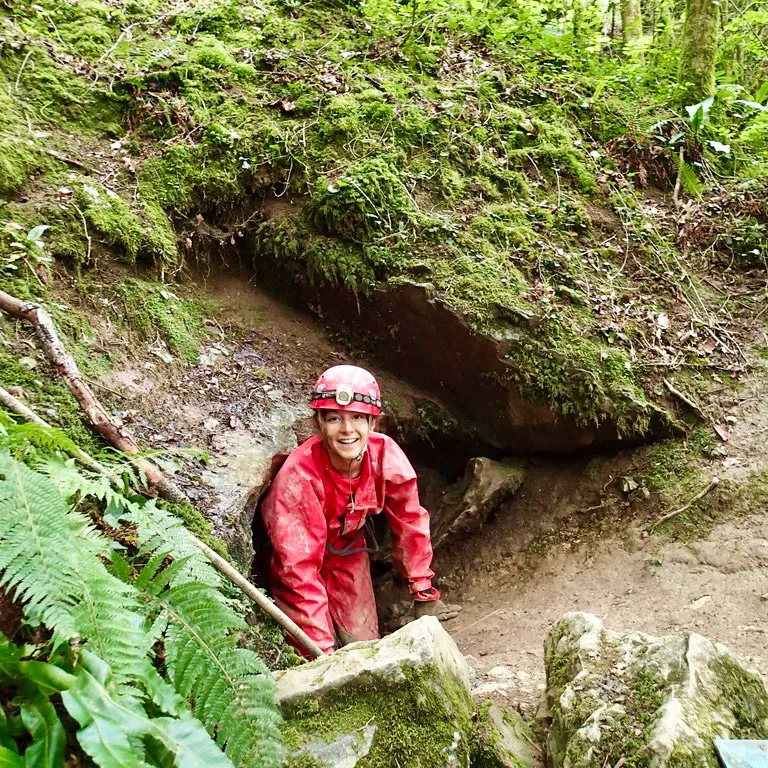 Spéléologue souriant en tenue rouge sortant d'une petite grotte couverte de mousse verte.