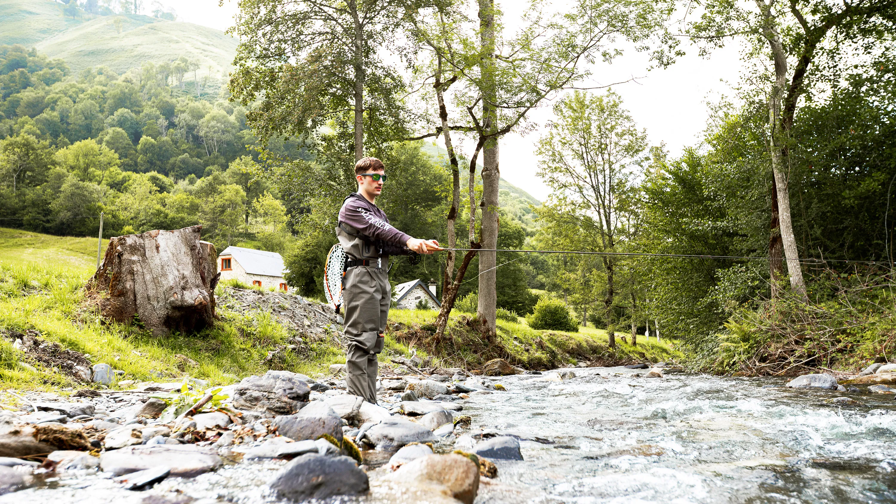 Un homme en waders pêche à la mouche dans une rivière entourée d'arbres et de collines.