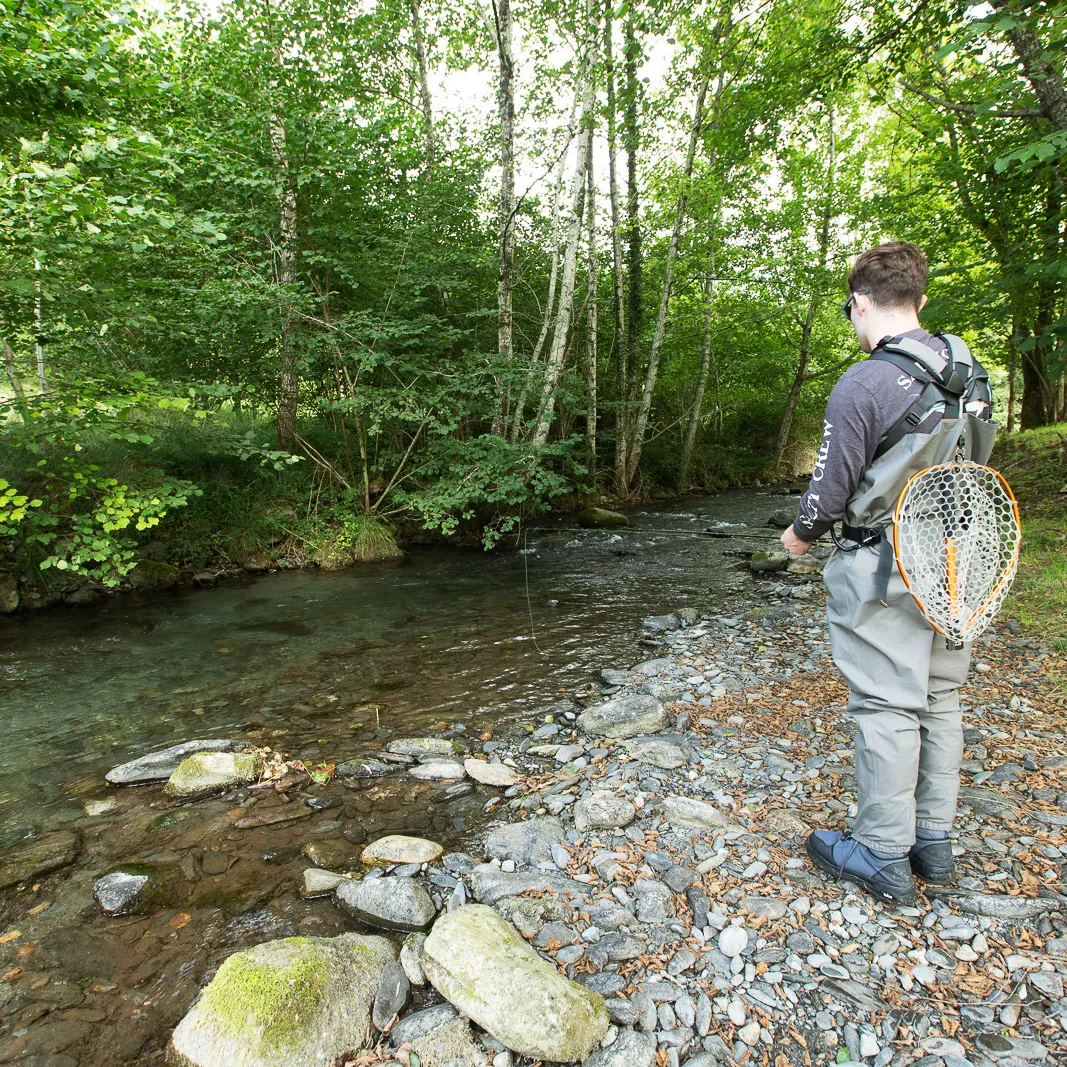 Un homme en waders pêche dans une rivière claire bordée d'arbres verts et de rochers.