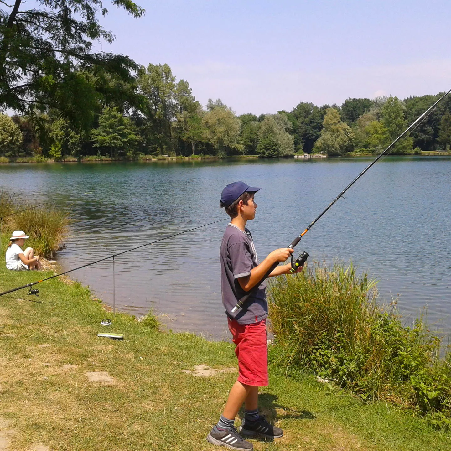 Un garçon pêche au bord d’un lac tandis qu’un enfant en chapeau est assis près de l’eau.