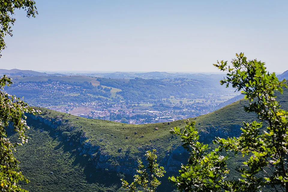 Vue sur une vallée avec des collines verdoyantes, des arbres et un village en arrière-plan.