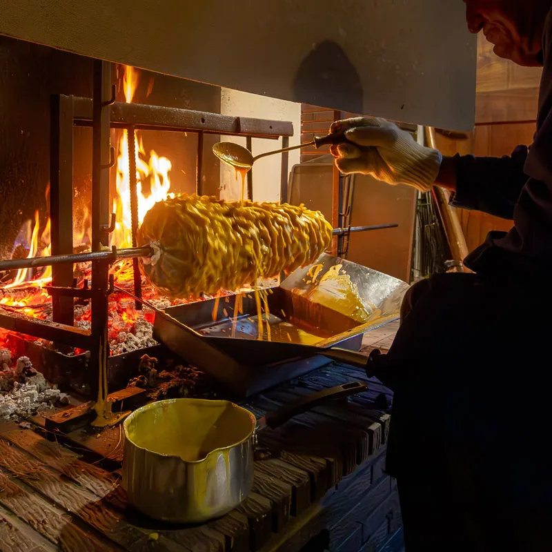 Un homme verse du mélange jaune fondu sur un gâteau en broche tournant près d'un feu.