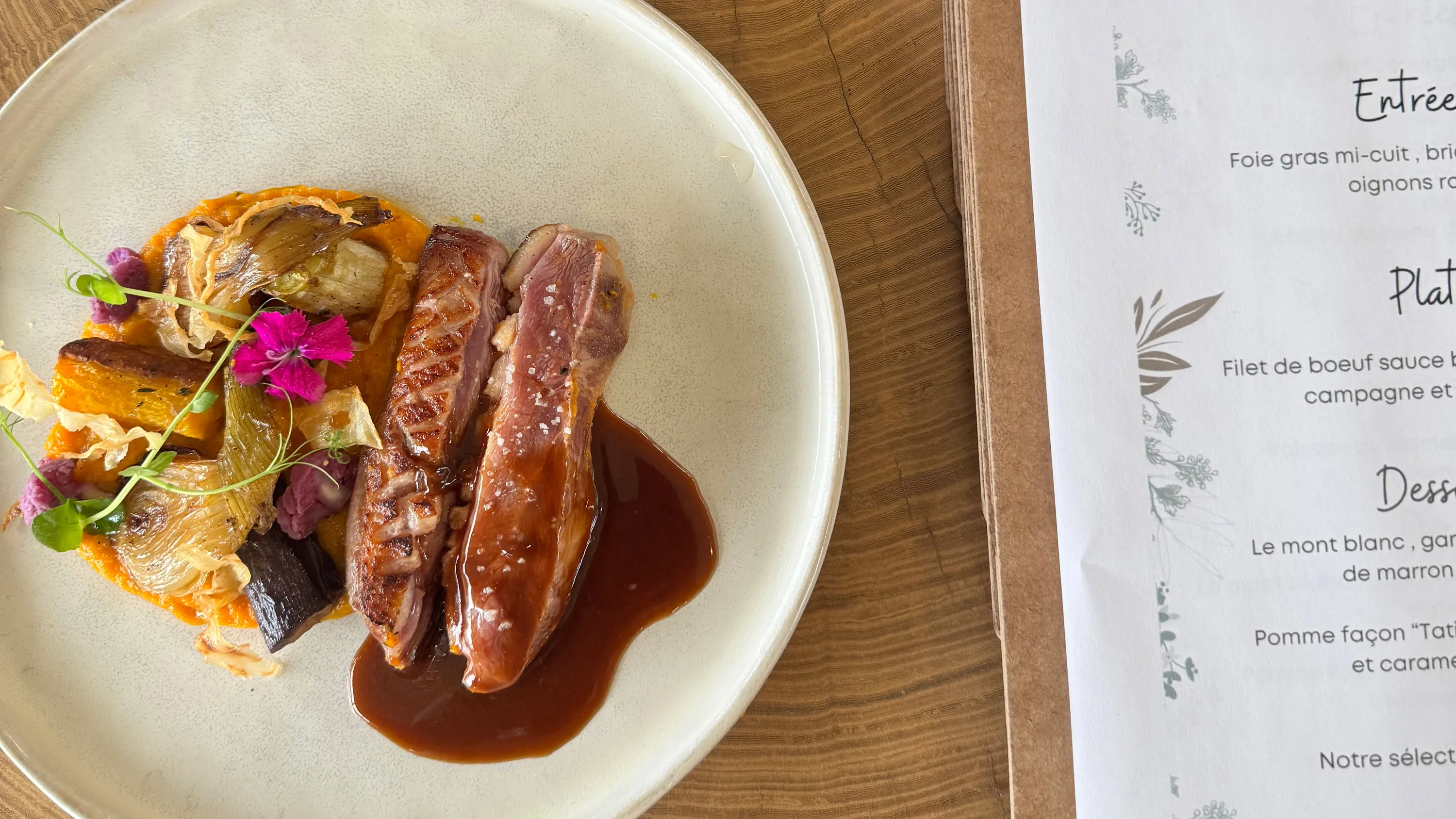 Assiette avec magret de canard en sauce, légumes grillés, et fleurs comestibles sur table en bois.