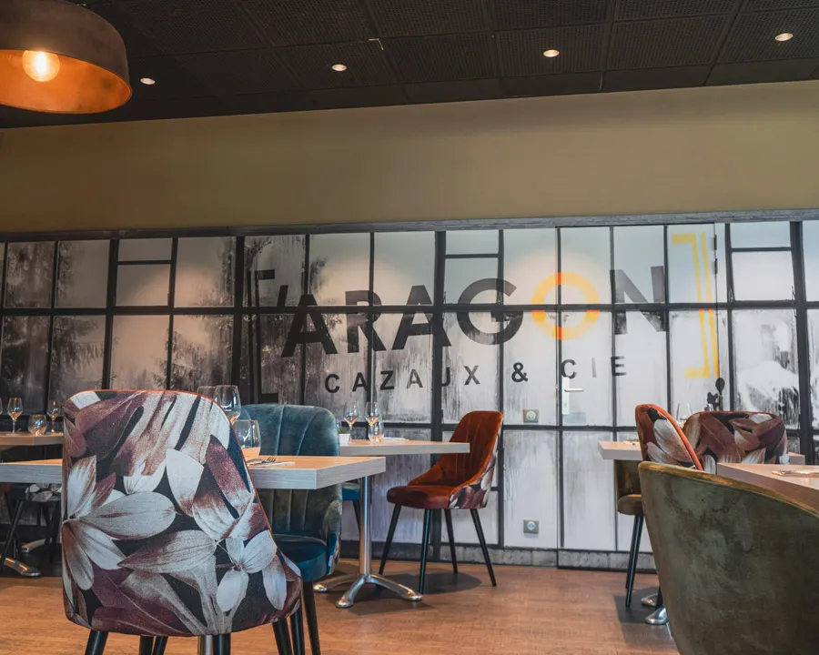 Intérieur moderne d'un restaurant avec chaises colorées et grandes fenêtres décorées du texte Aragon Cazaux & Cie.