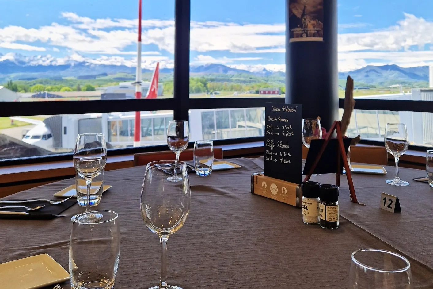 Table dressée avec verres et menu devant une fenêtre de restaurant face à un aéroport et montagnes.