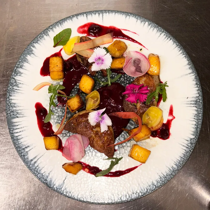 Assiette de foie poêlé décoré de sauce rouge, cubes de légumes, fleurs comestibles et feuilles vertes.