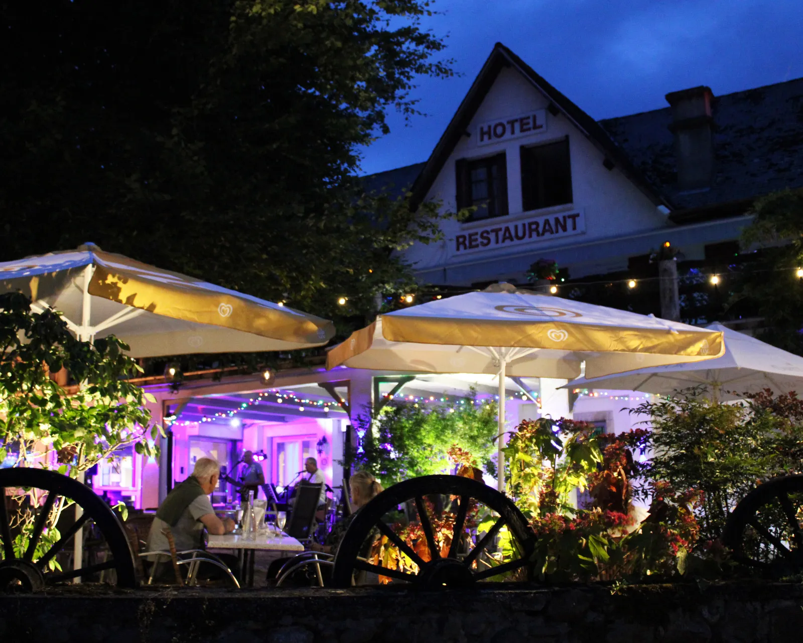 Terrasse éclairée au soir d'un hôtel-restaurant avec des clients et des parasols blancs.