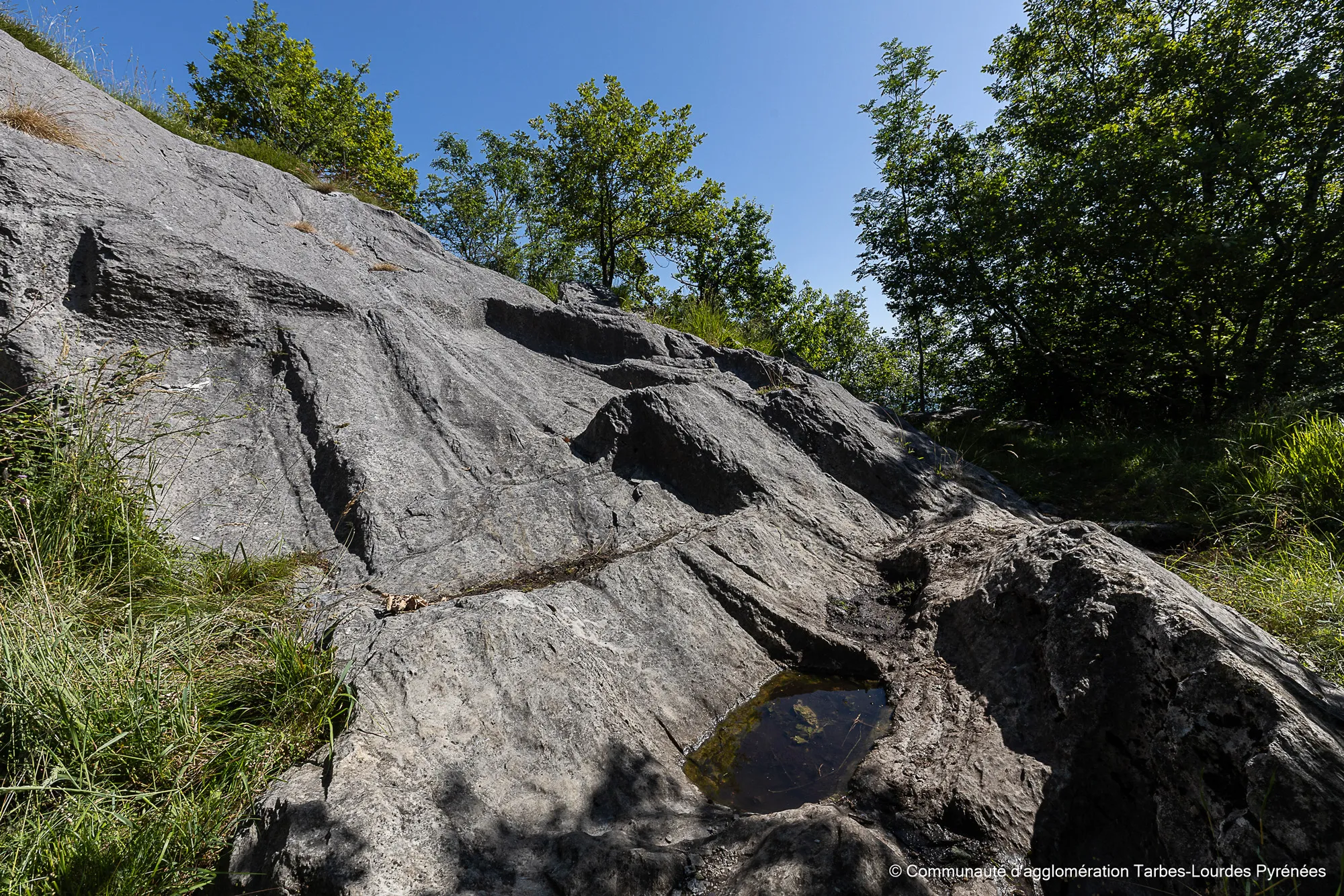 Vestiges de carrières avec traces de taille dans la roche grise sous un ciel bleu et des arbres verts.