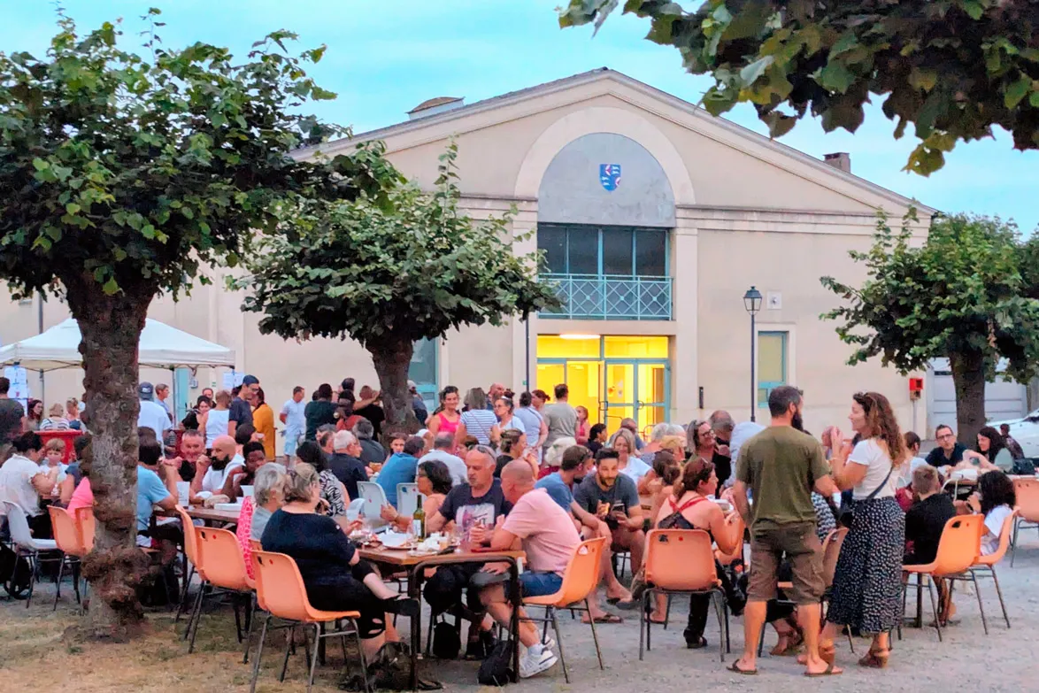 Personnes assises et debout autour de tables en extérieur lors d'un événement en soirée.