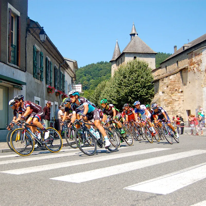 Cyclistes en course serrée dans un village avec un château et des spectateurs, ciel bleu clair.